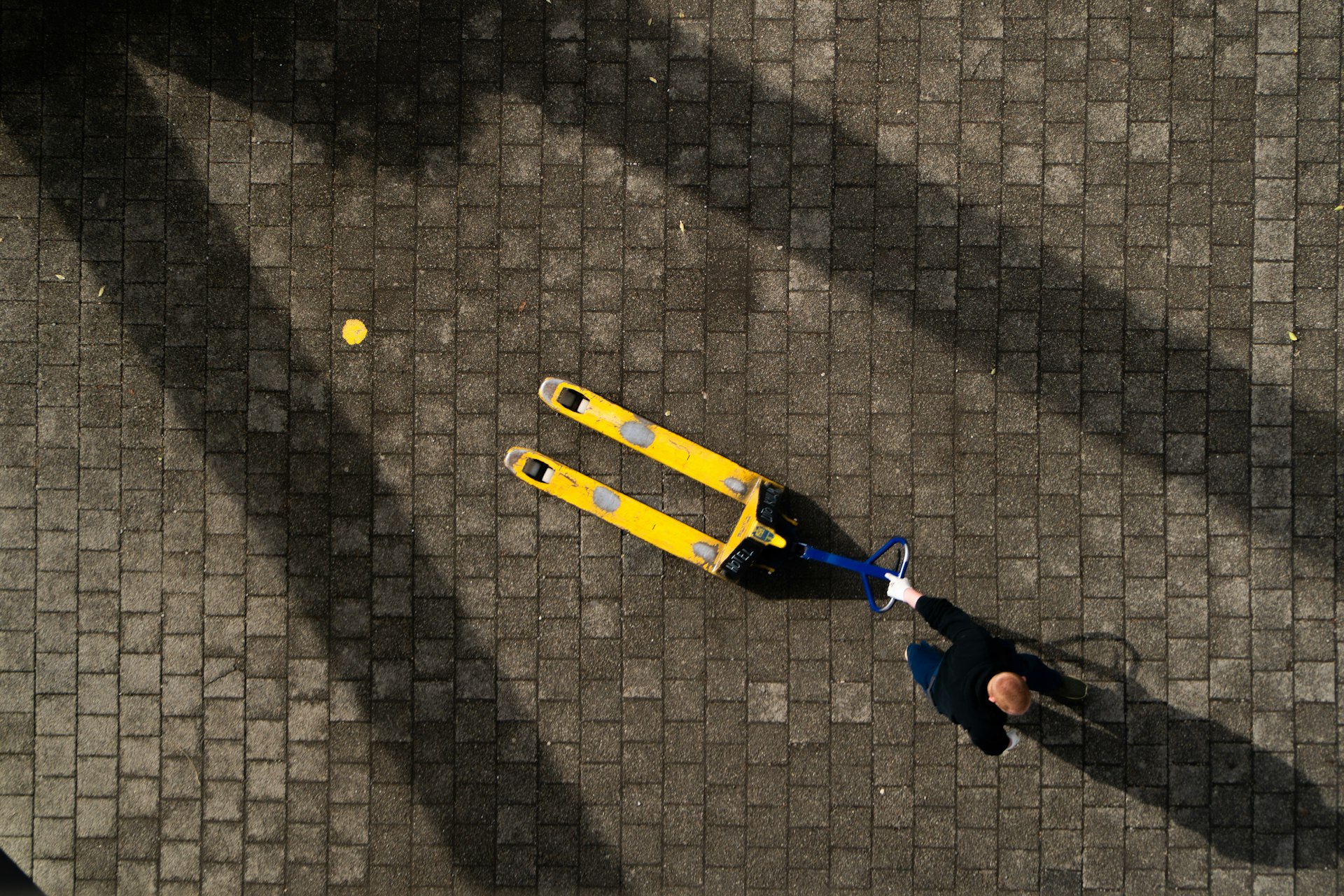 A worker seen from above, pulling an empty yellow hand pallet truck across a paved yard in low sun