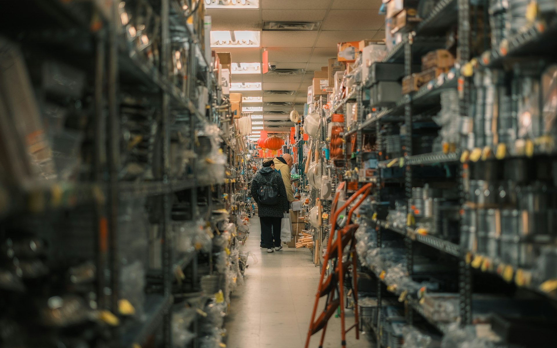 A narrow aisle in a packed catering-supply stockroom, steel shelving to the ceiling on both sides and two customers at the far end