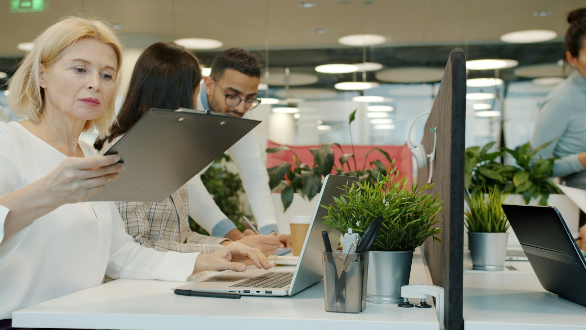 Colleagues at a shared desk in an open-plan office, one holding up a clipboard while another types on a laptop