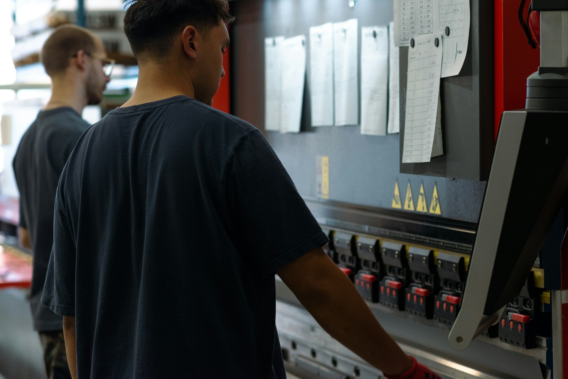 Two workers at a press brake, with printed job sheets clipped to the machine beside them