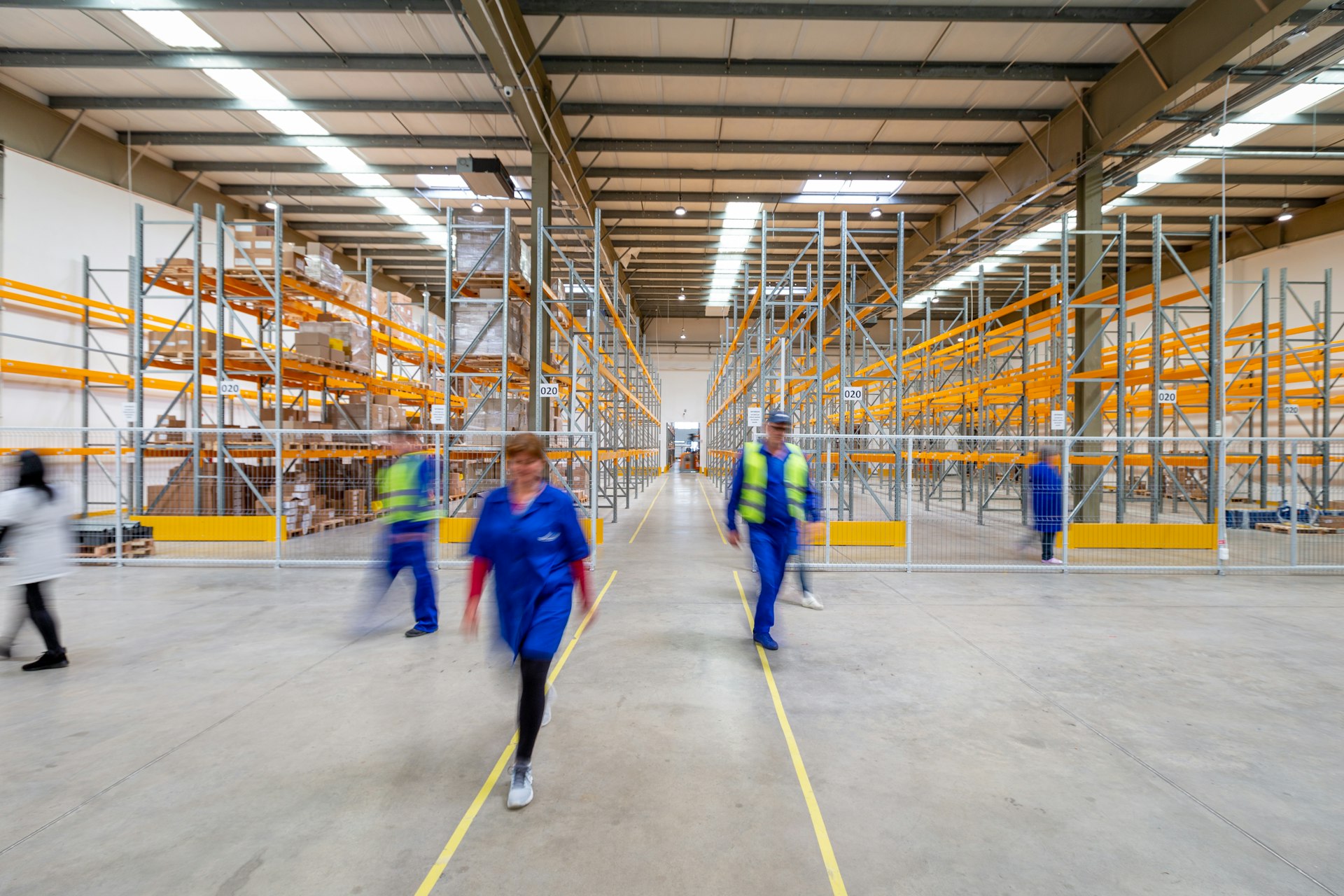 Warehouse staff crossing the central aisle of a distribution hall lined with orange pallet racking
