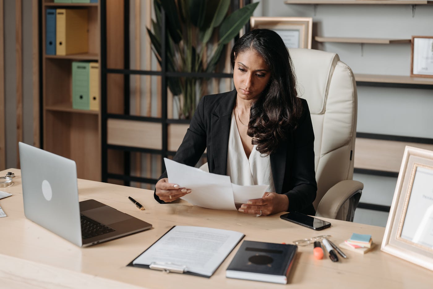 A professional at a desk reviewing printed documents, laptop and notebook beside her